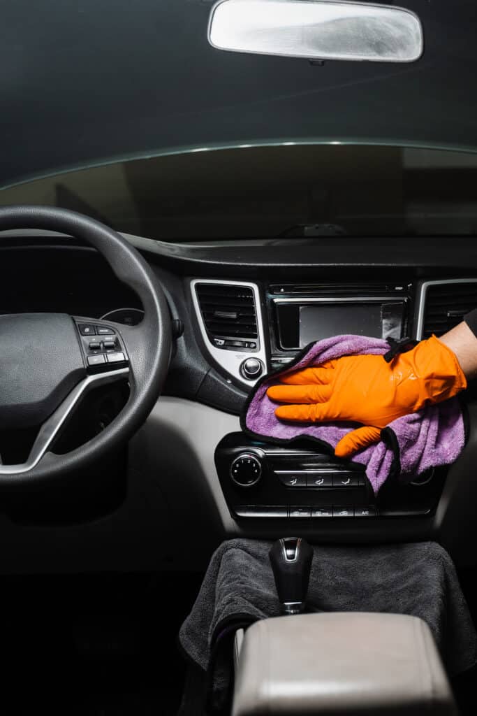 Detailing auto: main en gant orange nettoyant tableau de bord et console centrale avec chiffon microfibre violet.