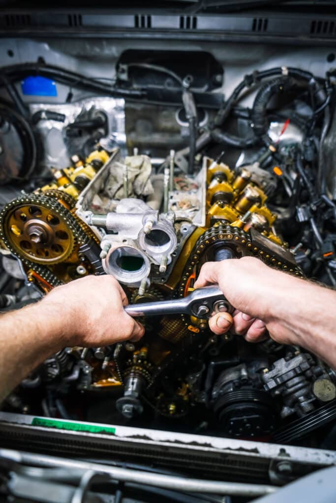 Mains de mécanicien serrant un boulon sur un moteur de voiture ouvert avec une clé à cliquet, chaînes de distribution visibles.